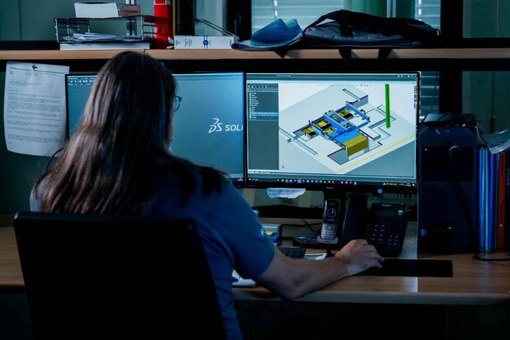 The image shows an operator interacting with CAD design software on a computer, used to develop industrial automation solutions On the screen a three-dimensional model of a production plant is visible, highlighting elements such as conveyor belts and actuators. The working environment is equipped with communication tools and documentation, suggesting a technical context oriented to the design of