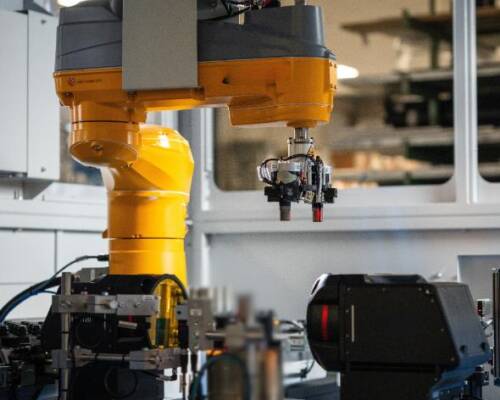 Robotic cell in an industrial plant featuring a yellow robotic arm equipped with a gripper for component handling, alongside sensors and inspection units typical of manufacturing applications
